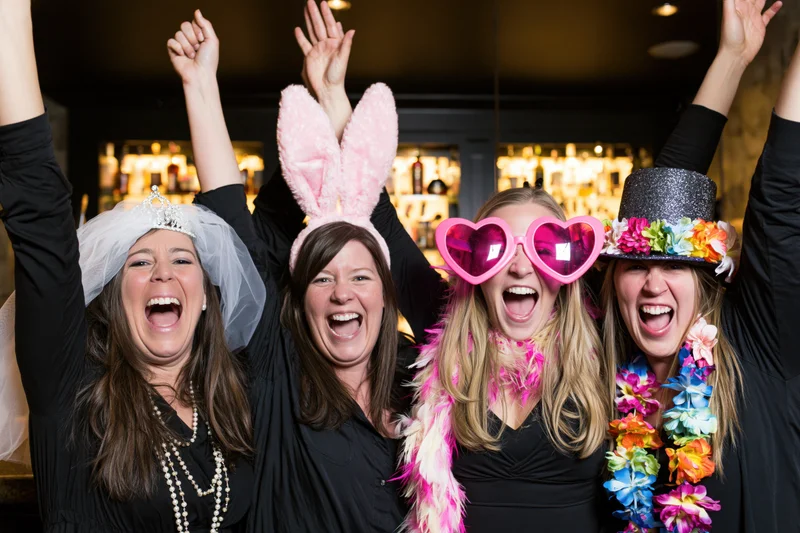 Four women laughing with arms raised at a nightclub party wearing festive accessories