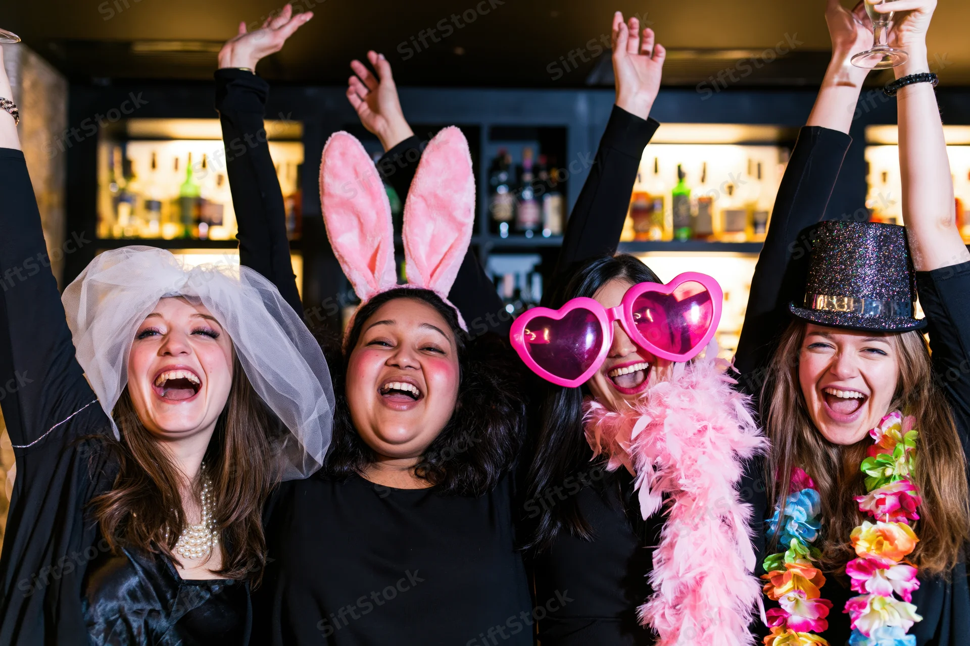 Four women laughing with arms raised at a nightclub party wearing festive costumes and accessories