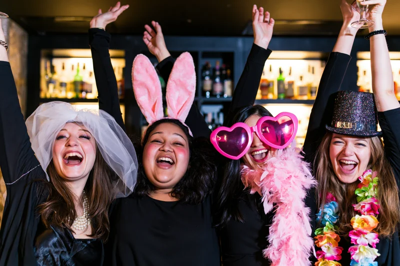 Four women laughing with arms raised at a nightclub party wearing festive costumes and accessories