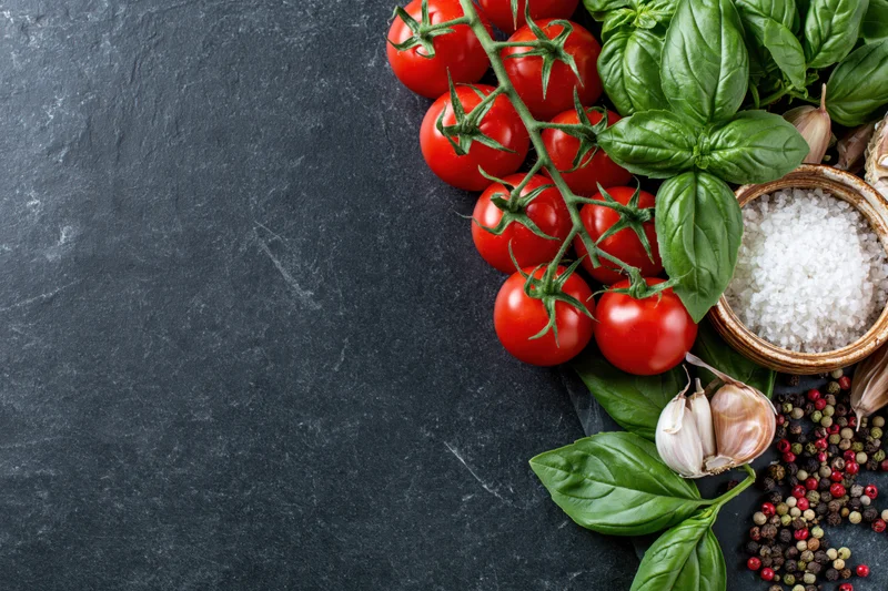 Fresh tomatoes, basil, garlic, salt and peppercorns arranged along the right edge of a dark slate surface