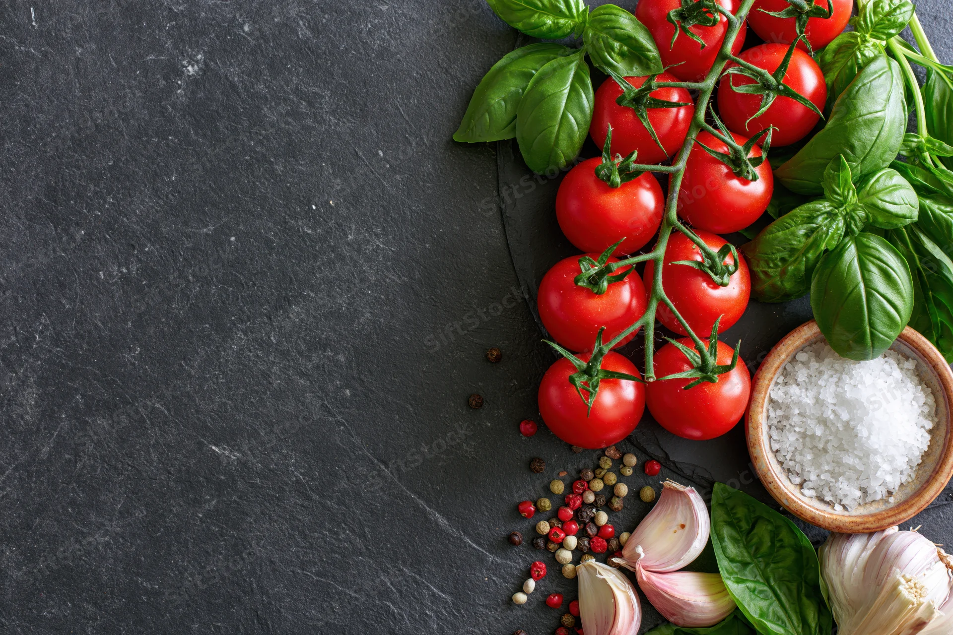 Fresh tomatoes on the vine with basil, garlic, peppercorns and salt arranged along the right edge of a dark slate surface