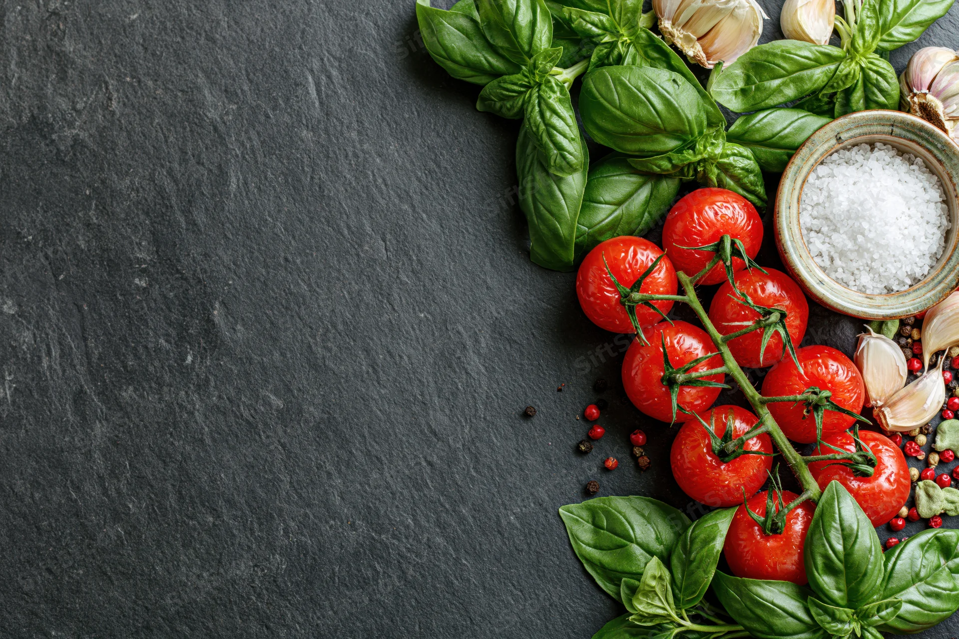 Fresh tomatoes, basil, garlic, and salt arranged along the right edge of a dark slate surface