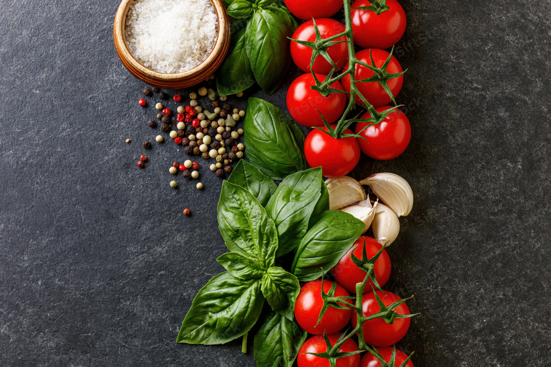 Fresh tomatoes basil garlic and peppercorns arranged along right edge on dark slate surface