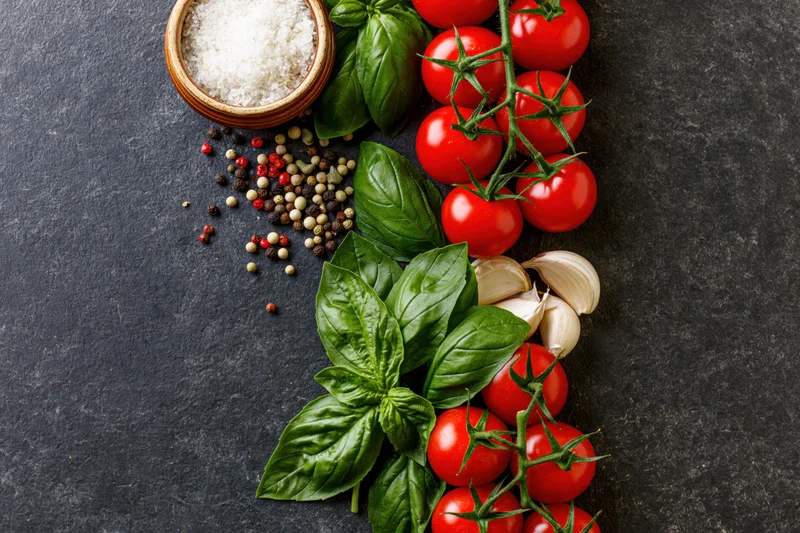 Fresh tomatoes basil garlic and peppercorns arranged along right edge on dark slate surface