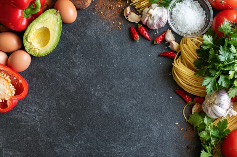 Fresh cooking ingredients arranged along the edges of a dark slate countertop with copyspace in center