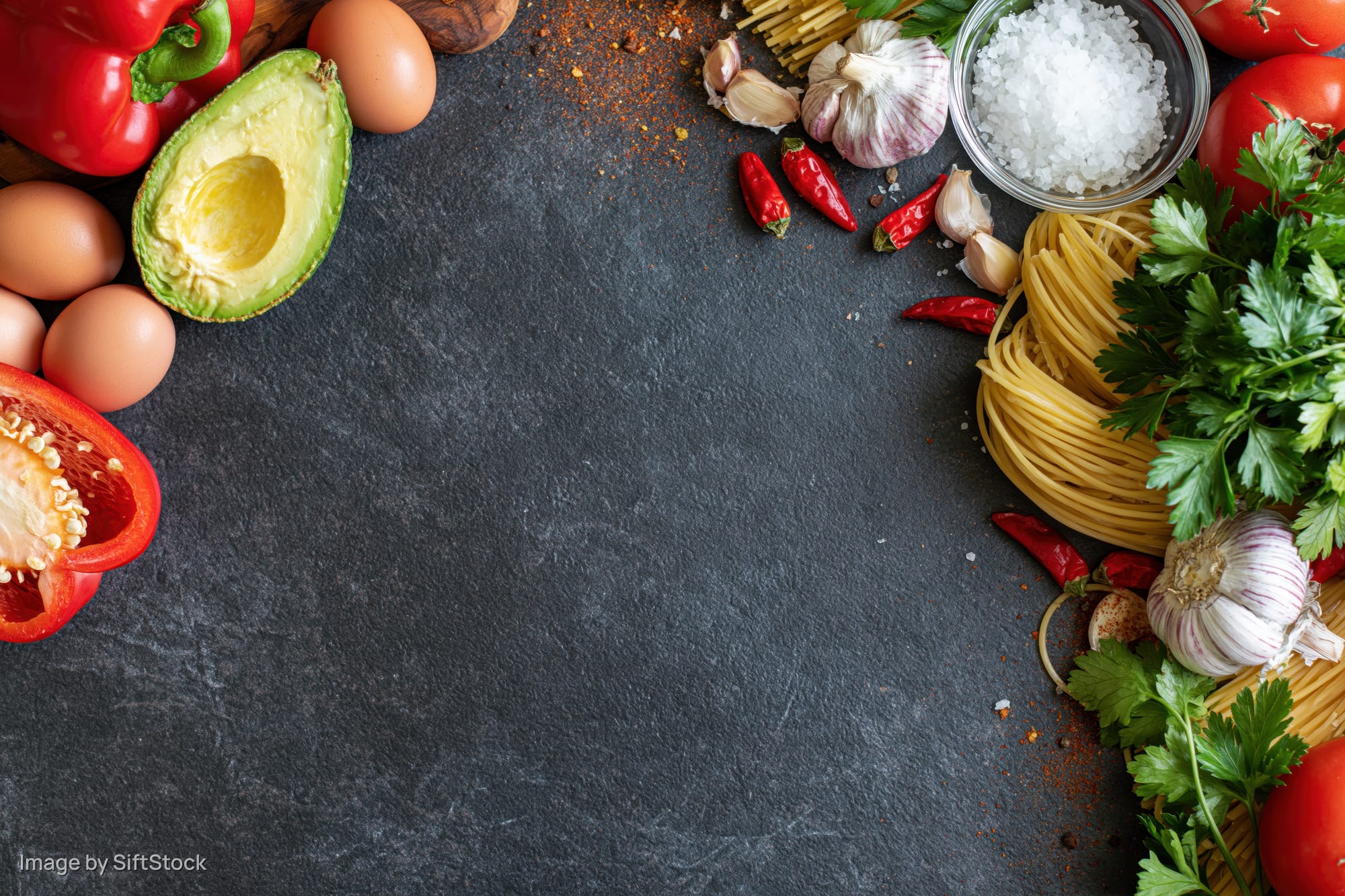 Fresh cooking ingredients arranged along the edges of a dark slate countertop with copyspace in center