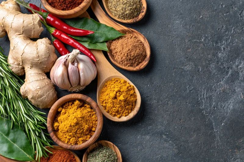Fresh ginger, garlic, rosemary, red chili, bay leaf, and various spices arranged on dark surface with copyspace