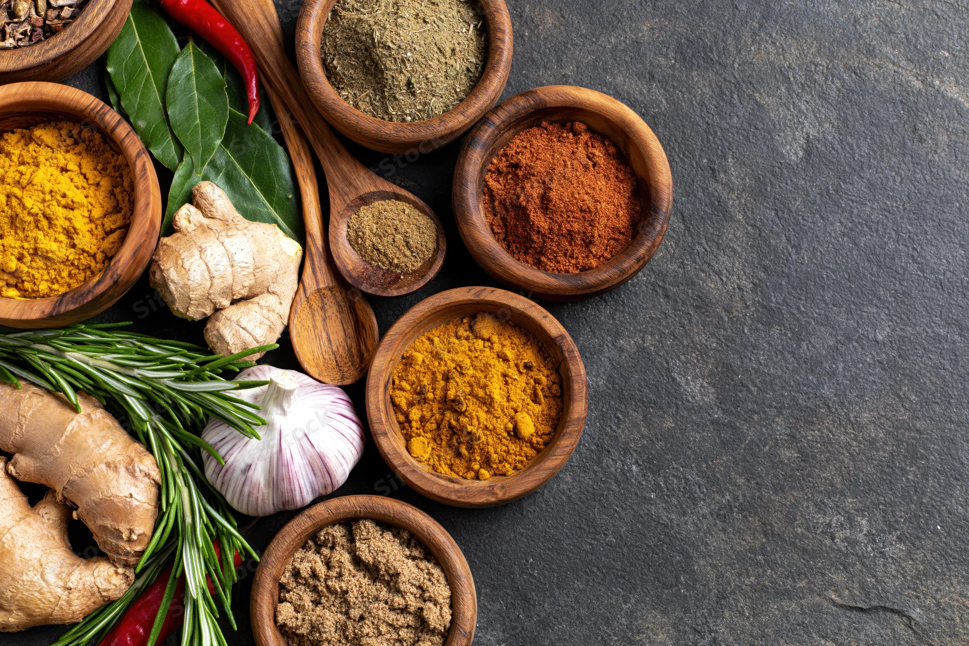 Various spices and herbs arranged in wooden bowls on a dark stone surface with copyspace
