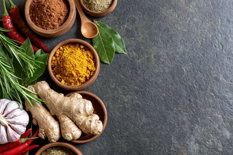 Fresh ginger, garlic, rosemary, red chili, bay leaf, and spices arranged in wooden bowls on a dark stone countertop