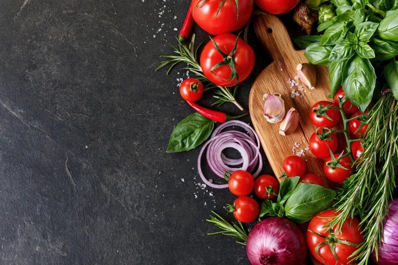 Fresh vegetables and herbs arranged along the right edge of a dark slate surface with copyspace on the left