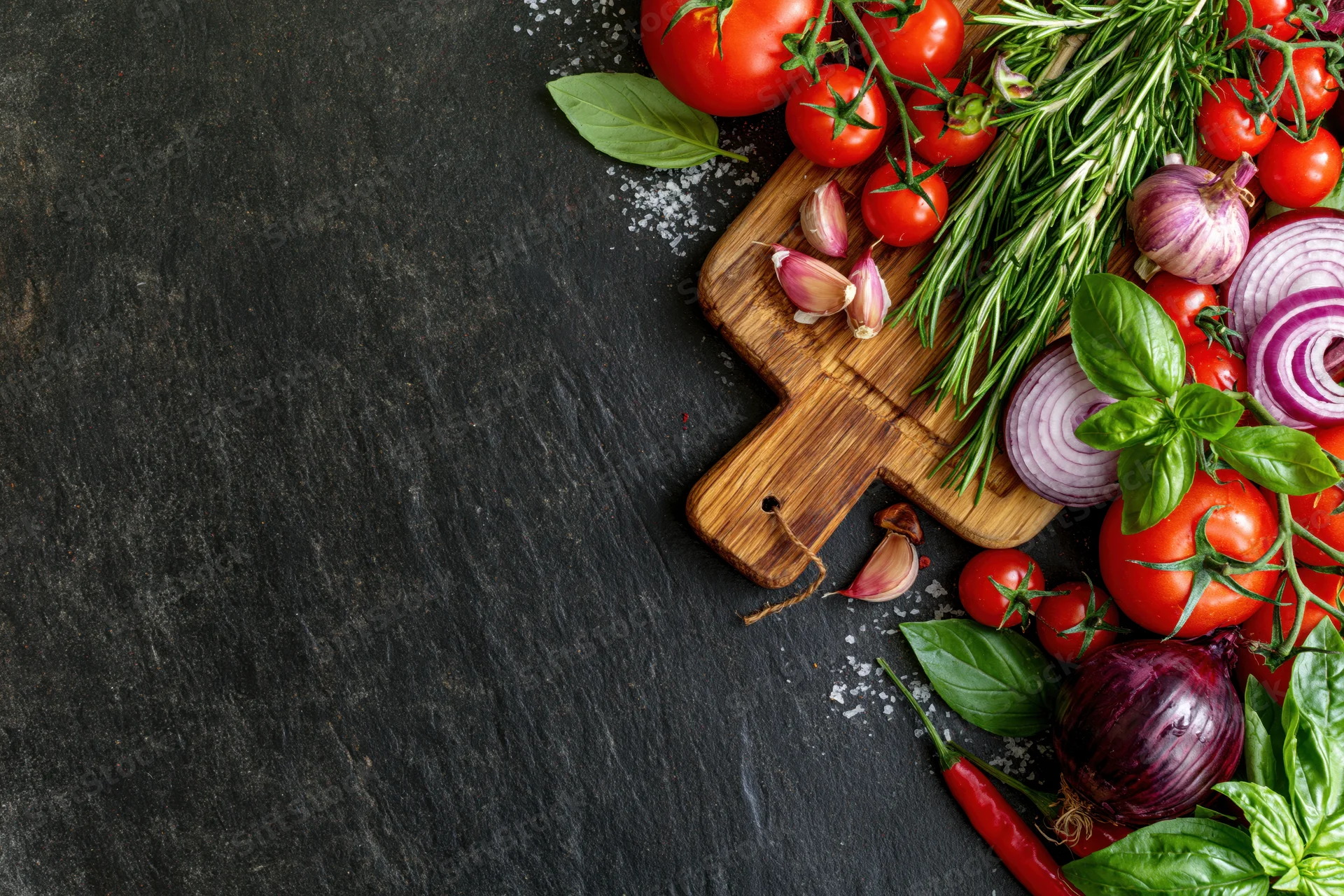 Fresh vegetables and herbs arranged on a wooden cutting board along the right edge of a dark slate surface
