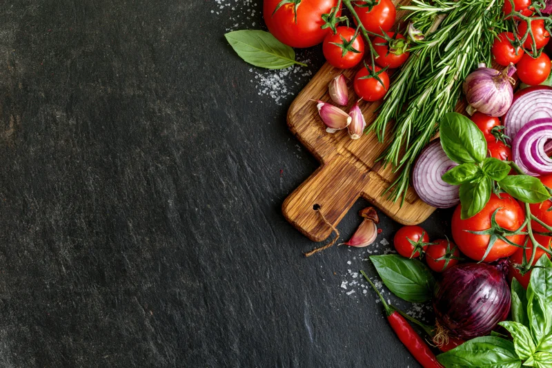 Fresh vegetables and herbs arranged on a wooden cutting board along the right edge of a dark slate surface