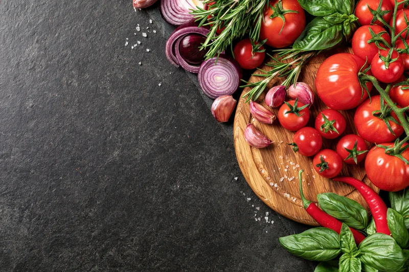 Fresh vegetables and herbs arranged on a wooden cutting board along the right edge of a dark slate surface