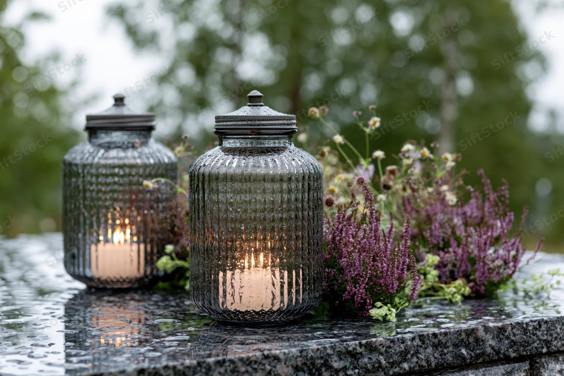 Two glass lanterns with lit candles on a wet granite grave marker surrounded by purple heather and wildflowers