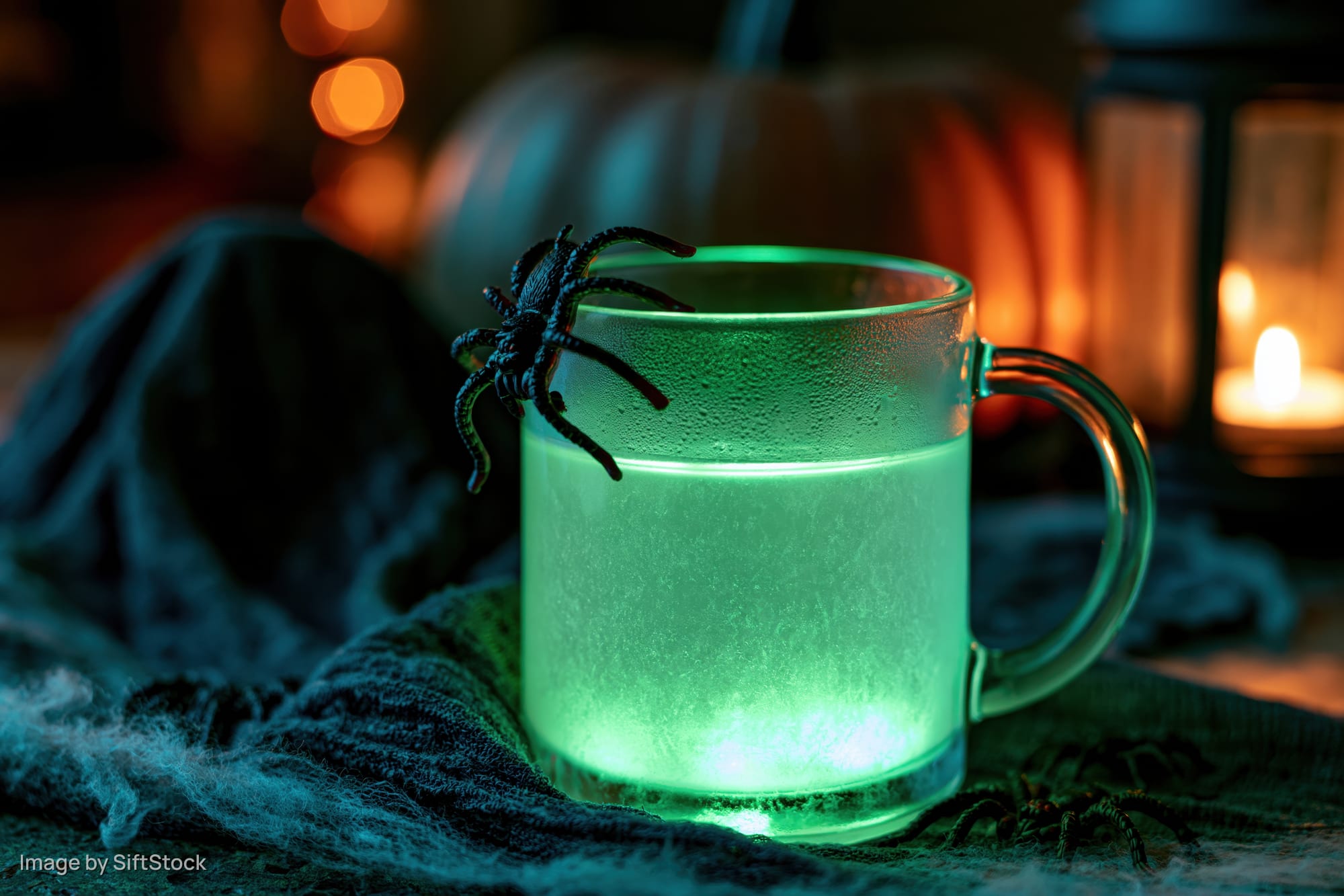 Glowing green drink in a glass mug with a fake spider on the rim, set on a dark fabric with Halloween decorations in the background
