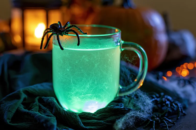 Glowing green drink in a glass mug with a plastic spider on the rim, set on a dark cloth with Halloween decorations and candles in the background