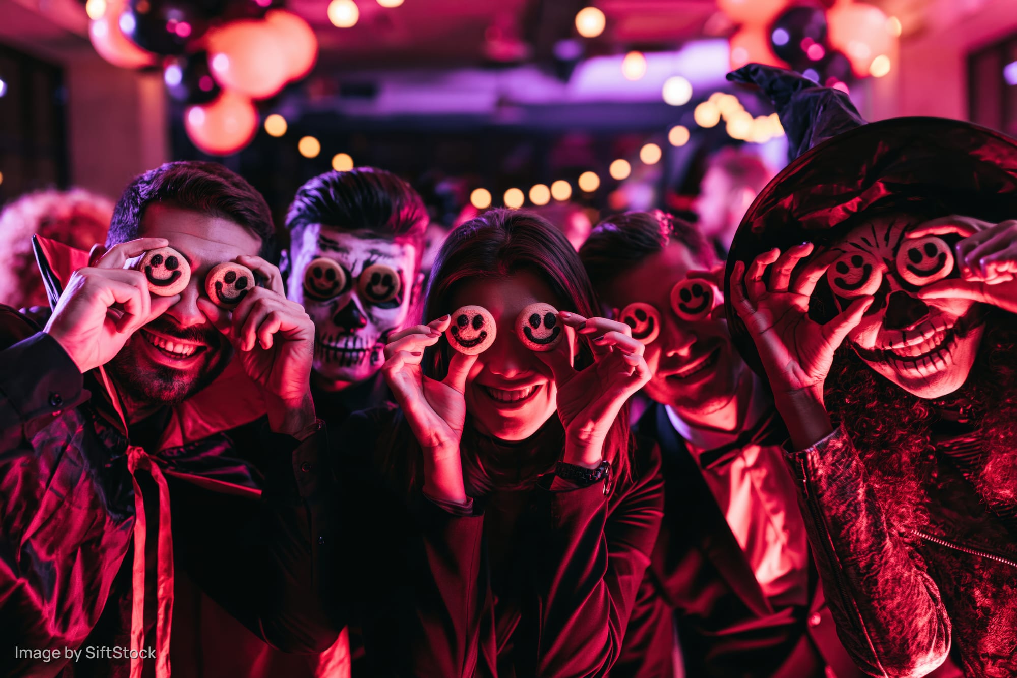 Five adults in halloween costumes holding smiley cookies over their eyes at a party