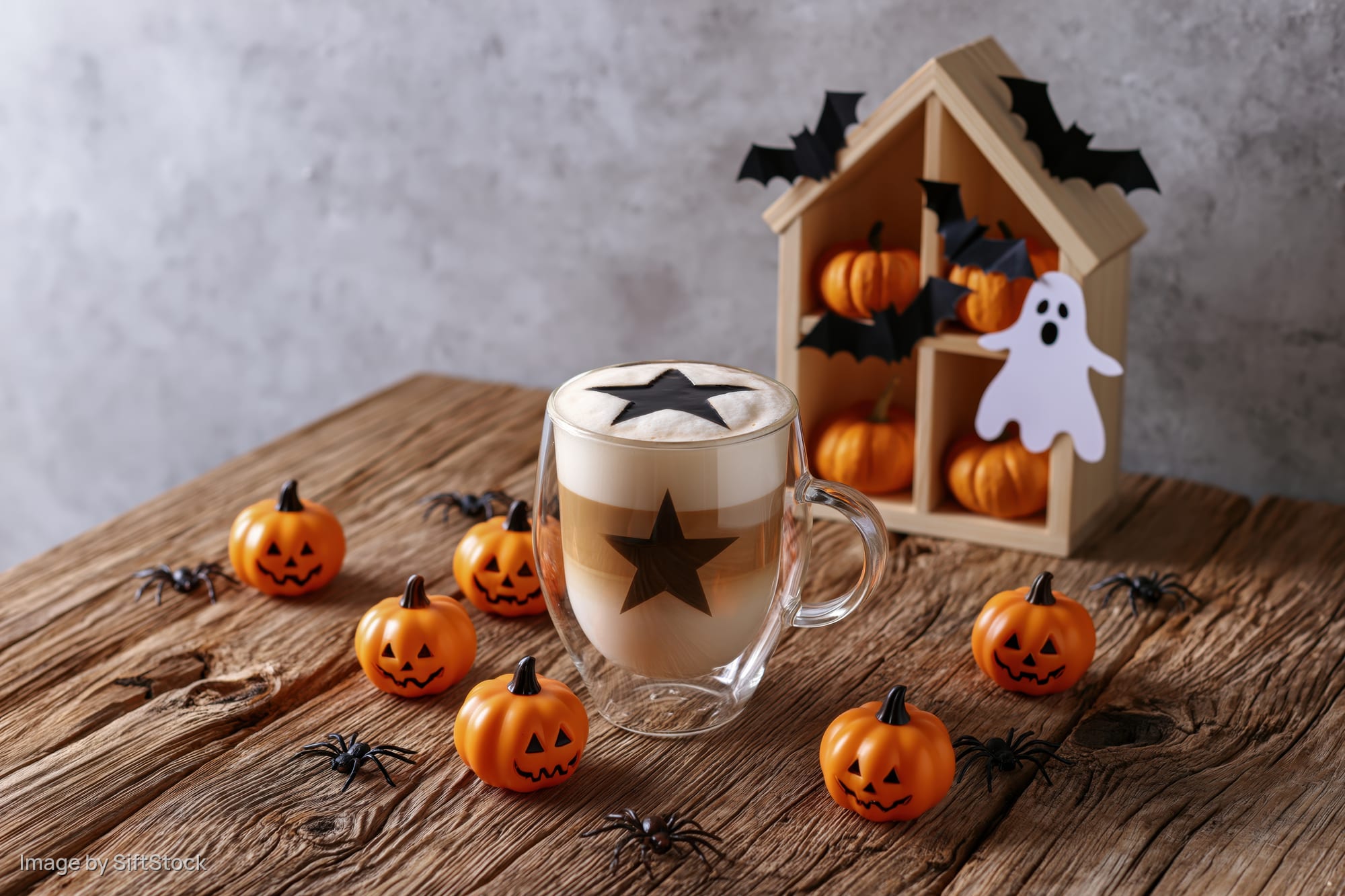 Halloween-themed coffee latte in a glass mug with star design on foam, surrounded by small pumpkins, spiders, ghost and bat decorations on a rustic wooden table