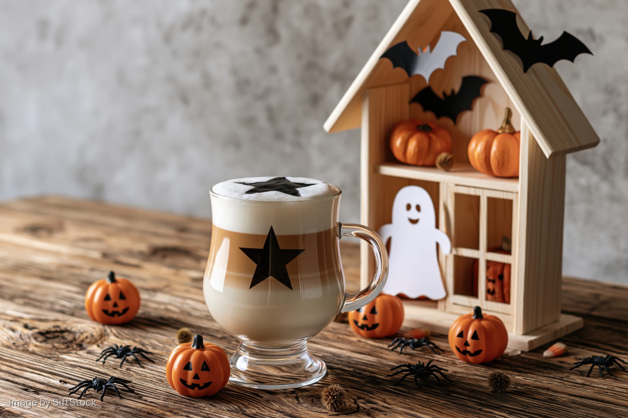 Halloween-themed latte in a glass mug with star foam design on rustic wooden table surrounded by pumpkins, spiders, and a decorated wooden house