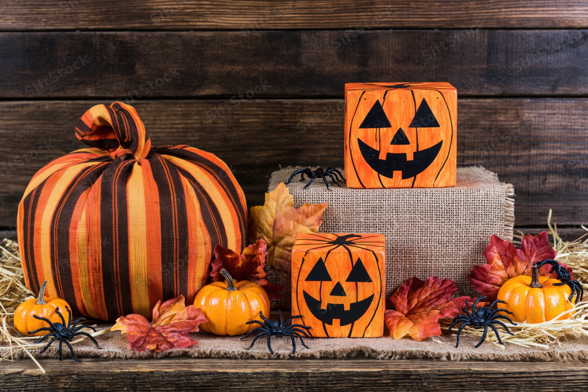 Halloween decorations including fabric pumpkins, wooden jack-o-lantern blocks, spiders, and autumn leaves arranged on a rustic wooden surface with hay and burlap