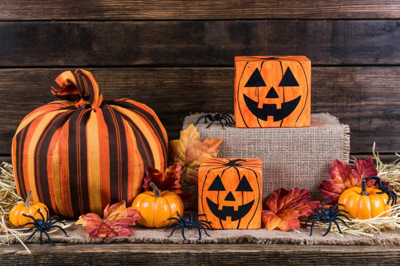 Halloween decorations including fabric pumpkins, wooden jack-o-lantern blocks, spiders, and autumn leaves arranged on a rustic wooden surface with hay and burlap