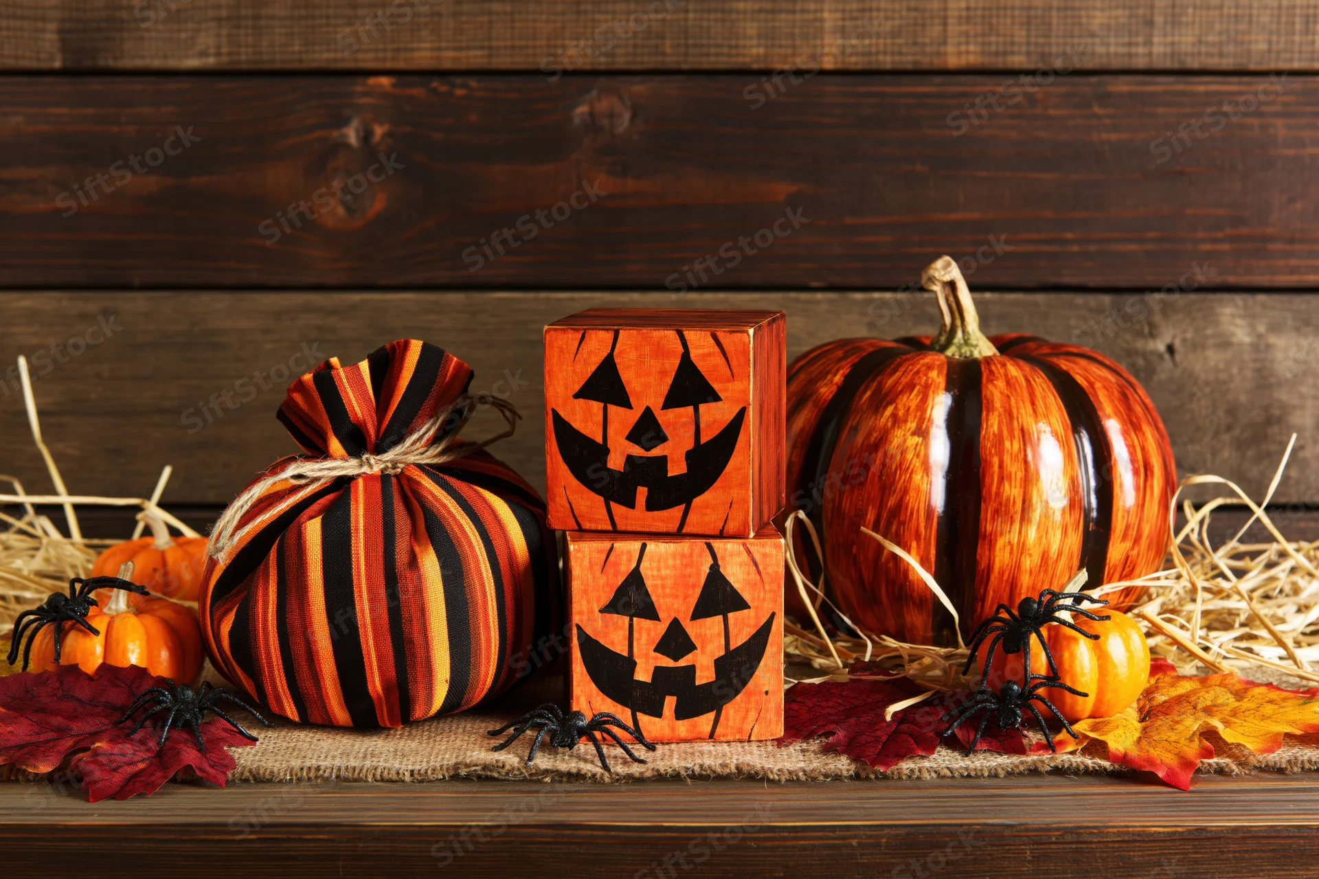 Halloween decorations including pumpkins, wooden jack-o-lantern blocks, and spiders arranged on a rustic wooden surface with hay and autumn leaves