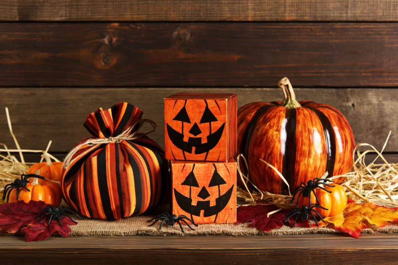Halloween decorations including pumpkins, wooden jack-o-lantern blocks, and spiders arranged on a rustic wooden surface with hay and autumn leaves
