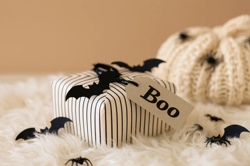 Halloween gift box wrapped in striped paper with a kraft tag reading boo on a fluffy white rug beside a knitted pumpkin and scattered bats and spiders