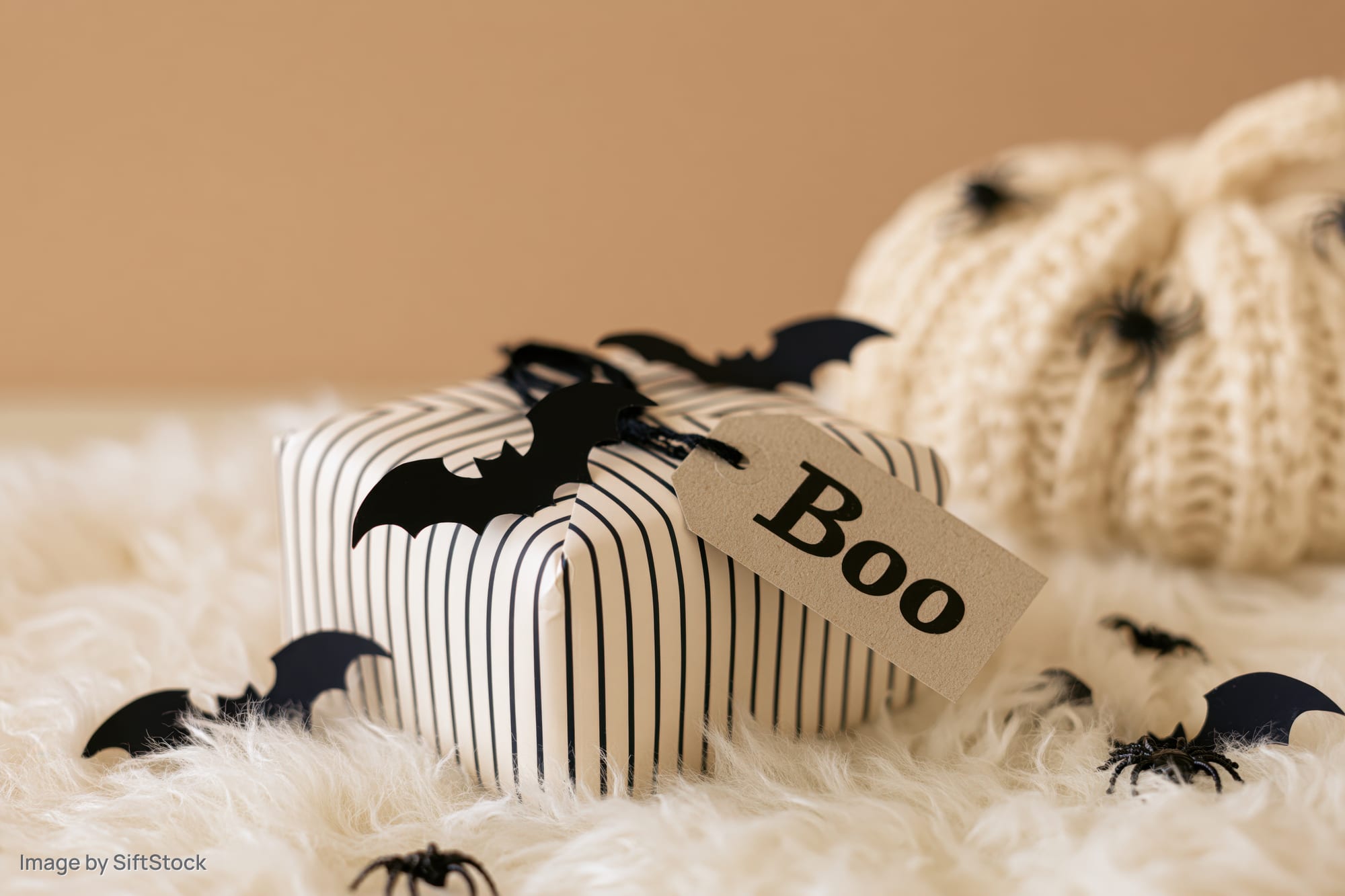 Halloween gift box wrapped in striped paper with a kraft tag reading boo on a fluffy white rug beside a knitted pumpkin and scattered bats and spiders