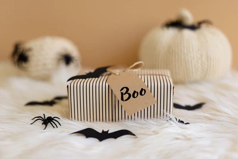 Halloween gift box wrapped in striped paper with a kraft tag reading boo on a white furry surface surrounded by bats and spiders and knitted pumpkins