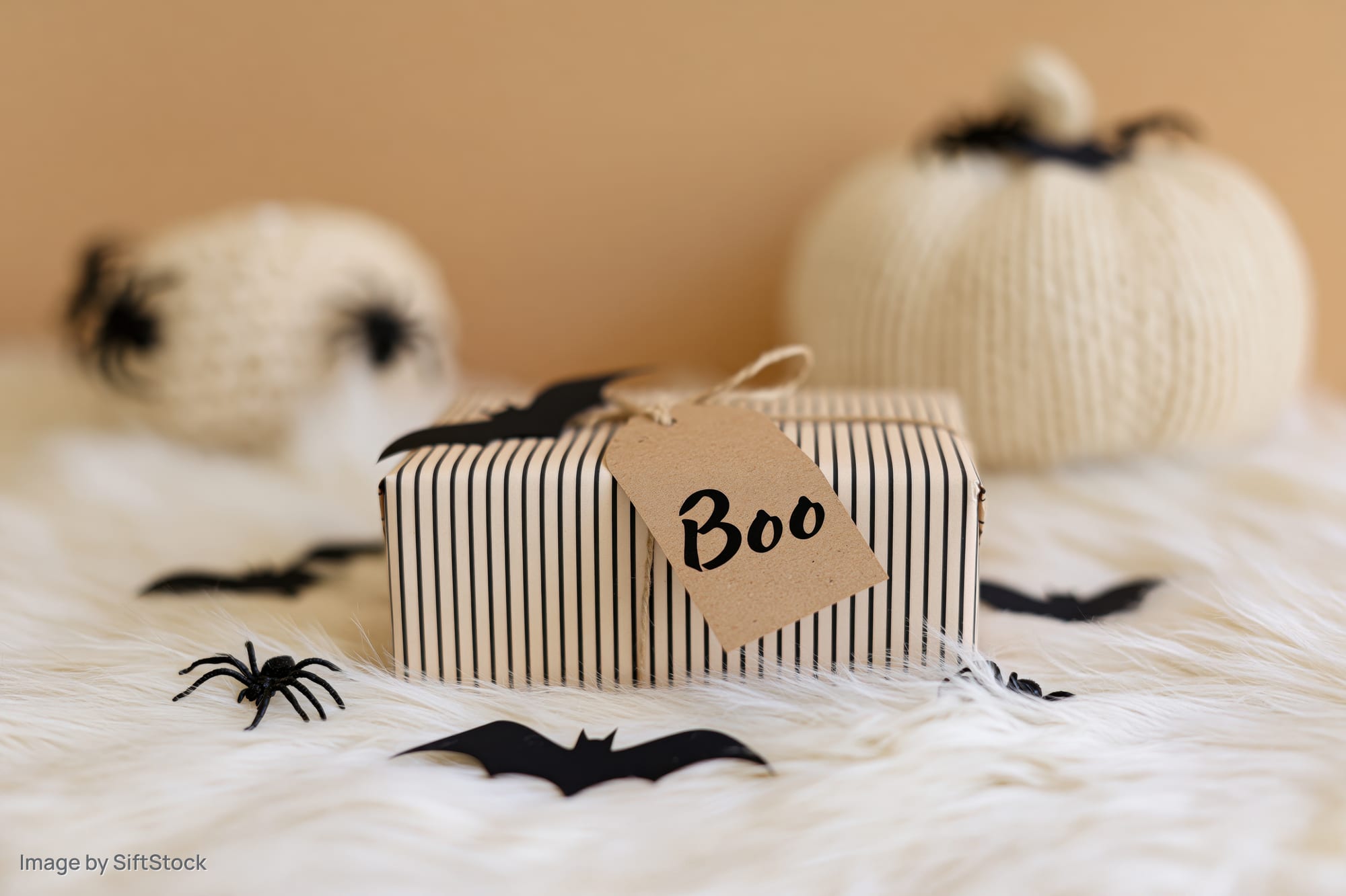 Halloween gift box wrapped in striped paper with a kraft tag reading boo on a white furry surface surrounded by bats and spiders and knitted pumpkins