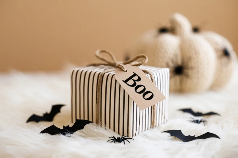 Halloween gift box wrapped in striped paper with a kraft tag reading boo on a white furry surface surrounded by bat and spider cutouts and a knitted pumpkin