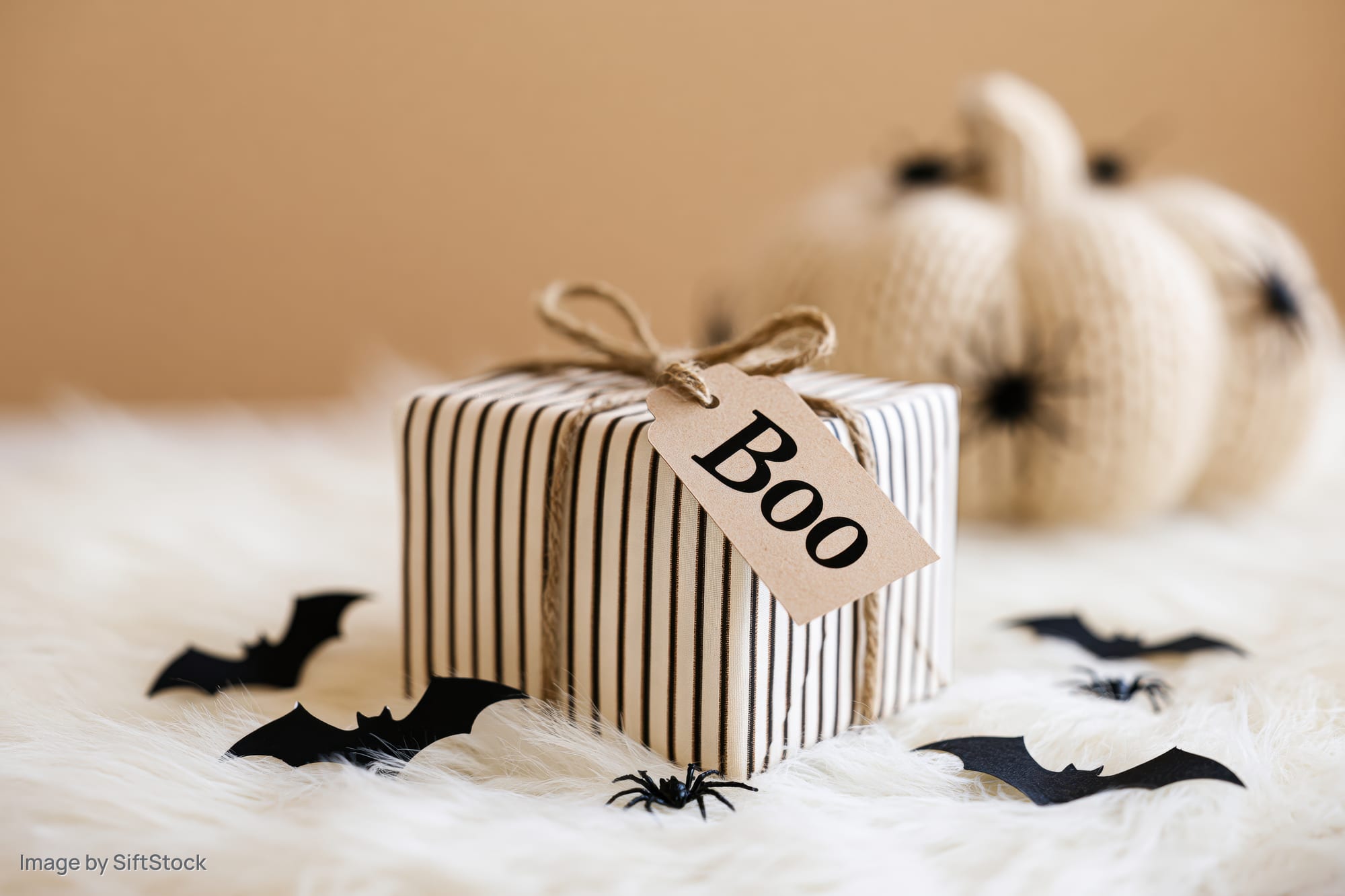 Halloween gift box wrapped in striped paper with a kraft tag reading boo on a white furry surface surrounded by bat and spider cutouts and a knitted pumpkin