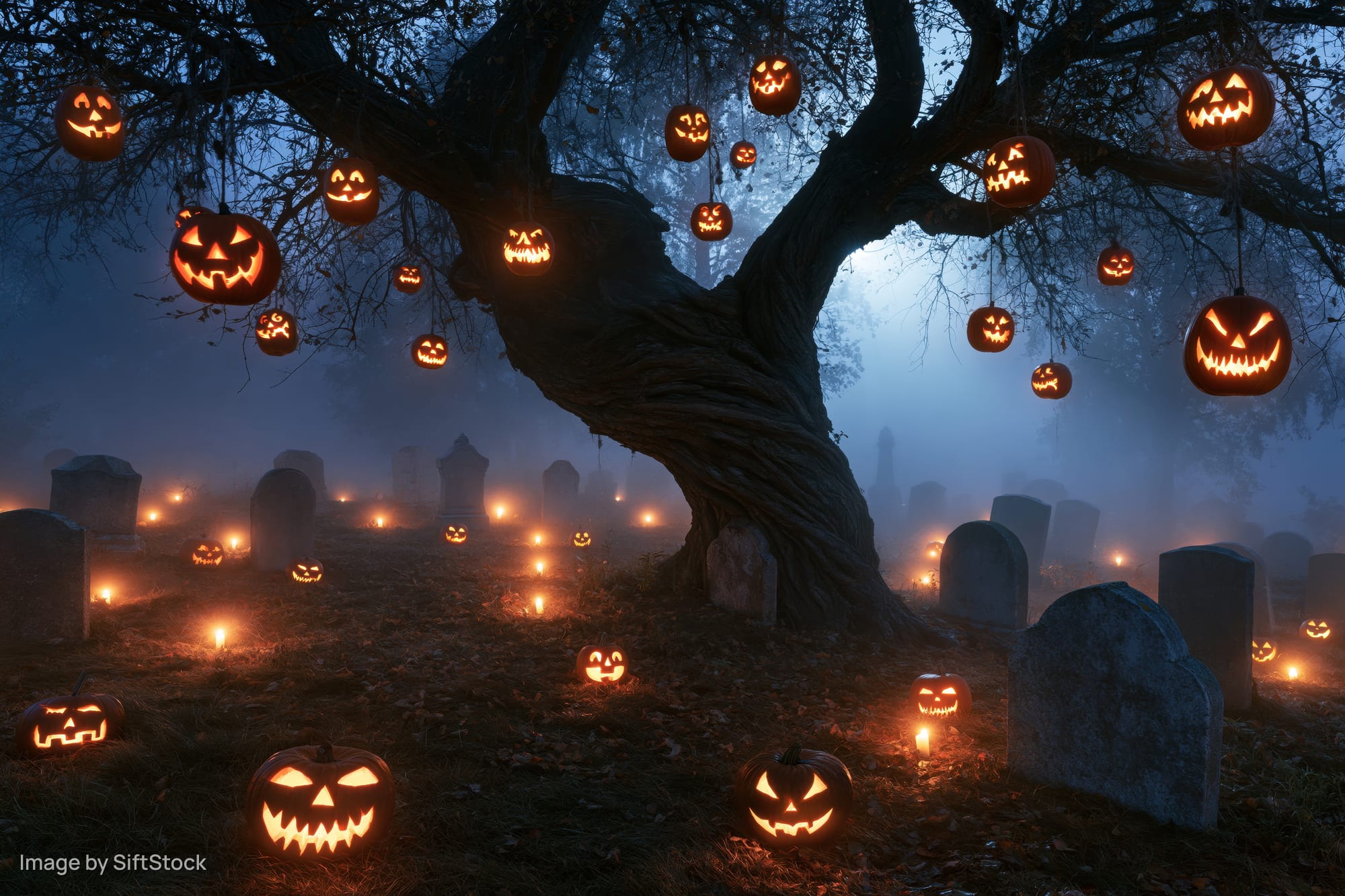 Carved jack-o-lanterns hanging from a gnarled tree in a foggy graveyard at night
