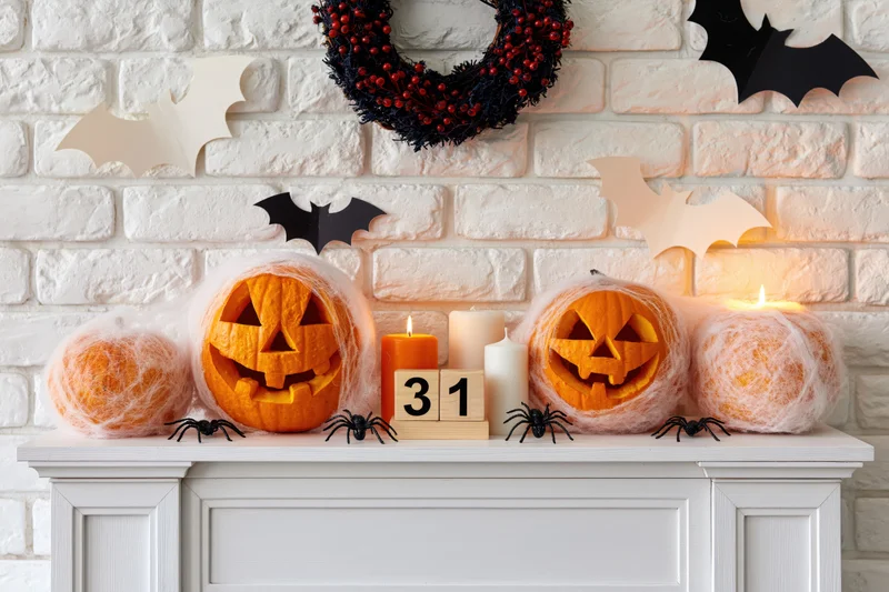 Carved jack-o-lanterns and candles on a white mantel with bats and wreath against a brick wall