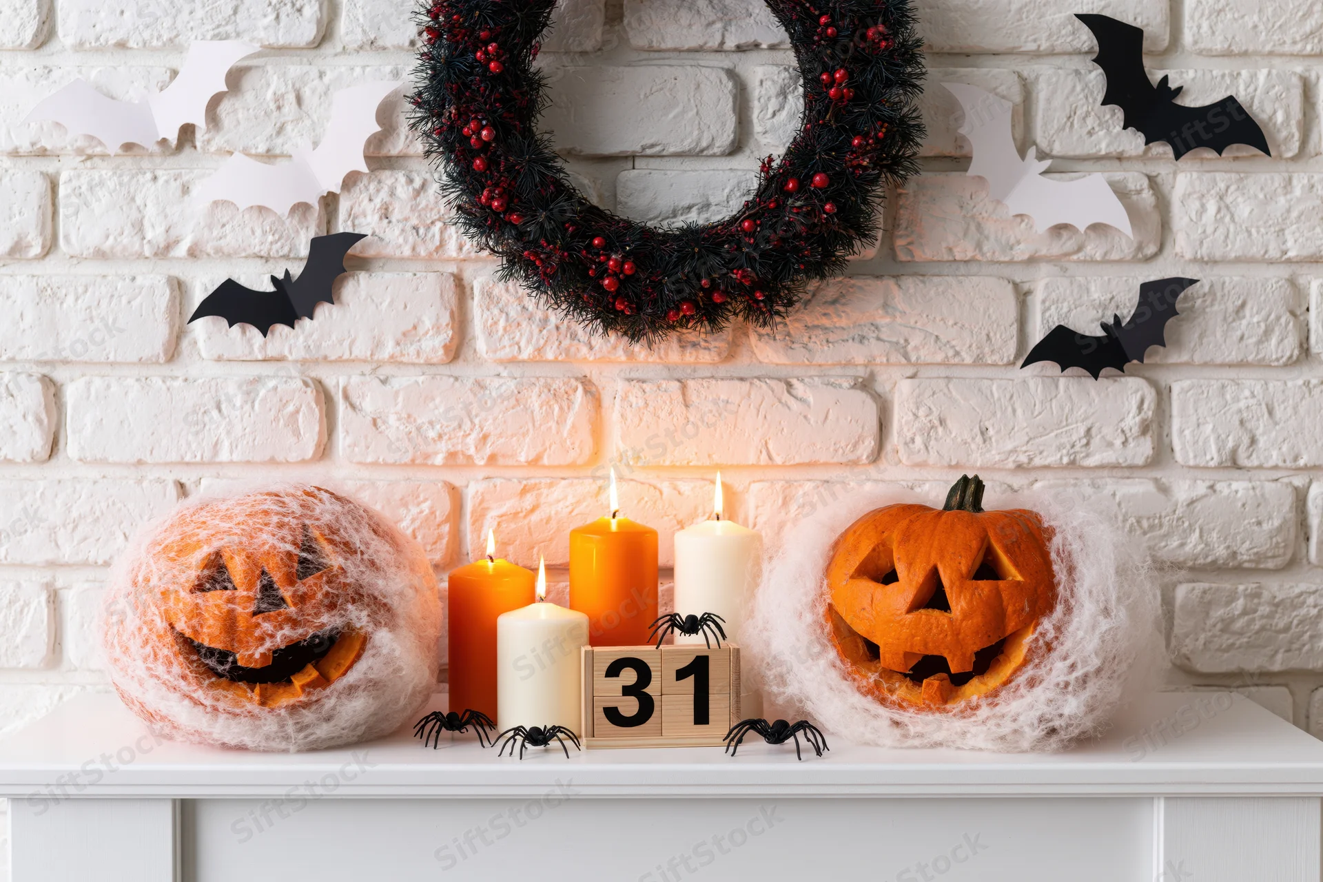 Carved jack-o-lanterns with candles and spiders on a mantel decorated with a wreath and bats against a brick wall