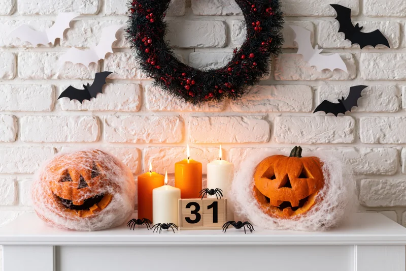 Carved jack-o-lanterns with candles and spiders on a mantel decorated with a wreath and bats against a brick wall
