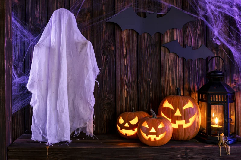 Halloween porch decoration with a white fabric ghost, carved pumpkins, and a lit lantern against a wooden wall with spiderwebs and bat cutouts