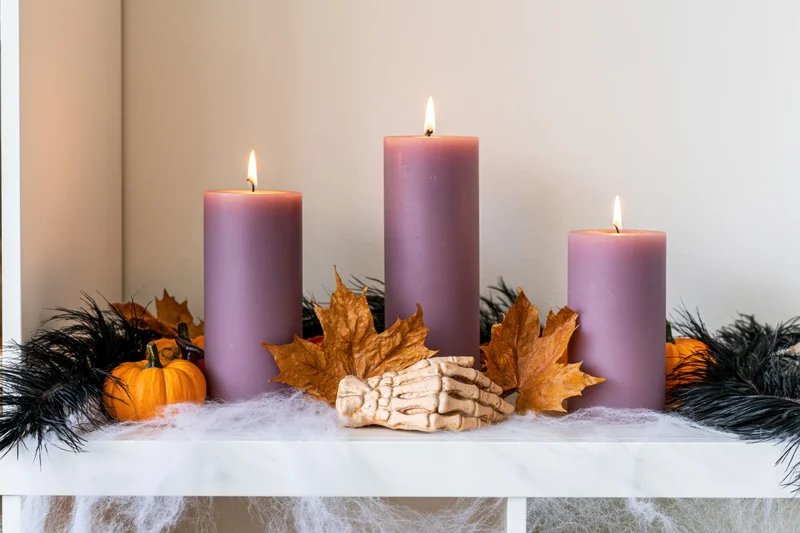 Three lit dusty purple pillar candles on a shelf with halloween decorations including pumpkins, skeleton hand, leaves, and cobwebs