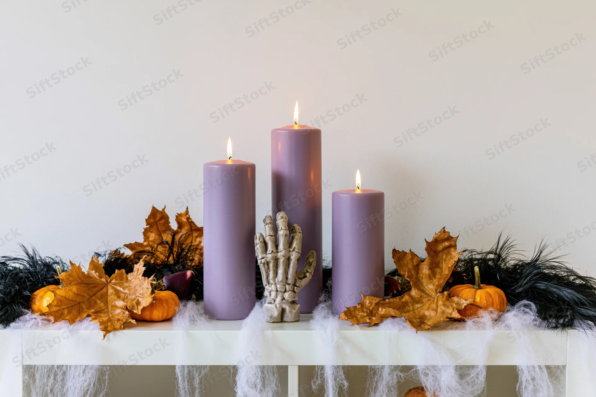 Halloween shelf decoration with lit purple candles, skeleton hand, pumpkins and autumn leaves arranged on a white table with cobweb draping