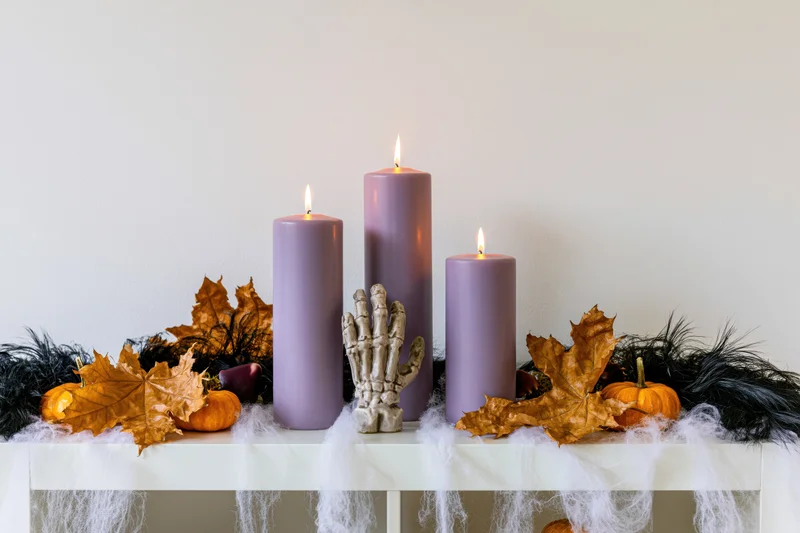 Halloween shelf decoration with lit purple candles, skeleton hand, pumpkins and autumn leaves arranged on a white table with cobweb draping