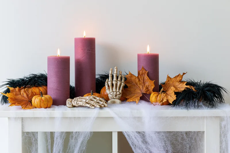 Halloween decoration with lit purple candles, pumpkins, skeleton hands and autumn leaves on a white shelf draped with spiderweb