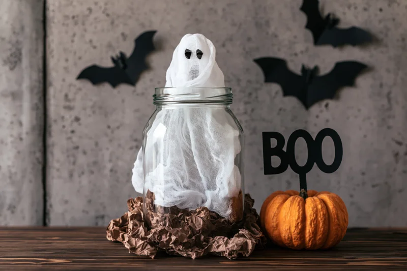 Ghost in a glass jar on a wooden table with a pumpkin and bat cutouts for halloween decoration