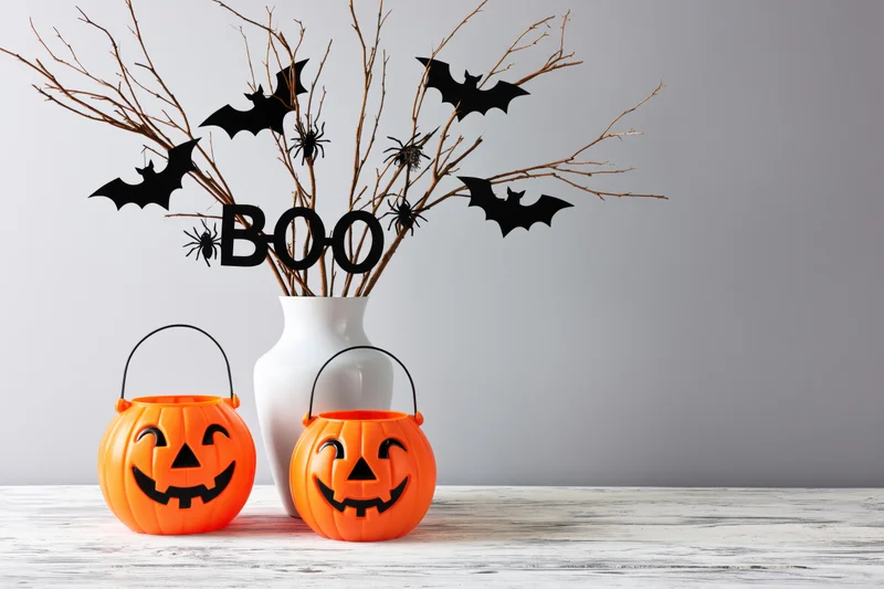 Two orange plastic jack-o-lantern buckets and a white vase with dry branches, bats, spiders and a boo sign on a wooden tabletop