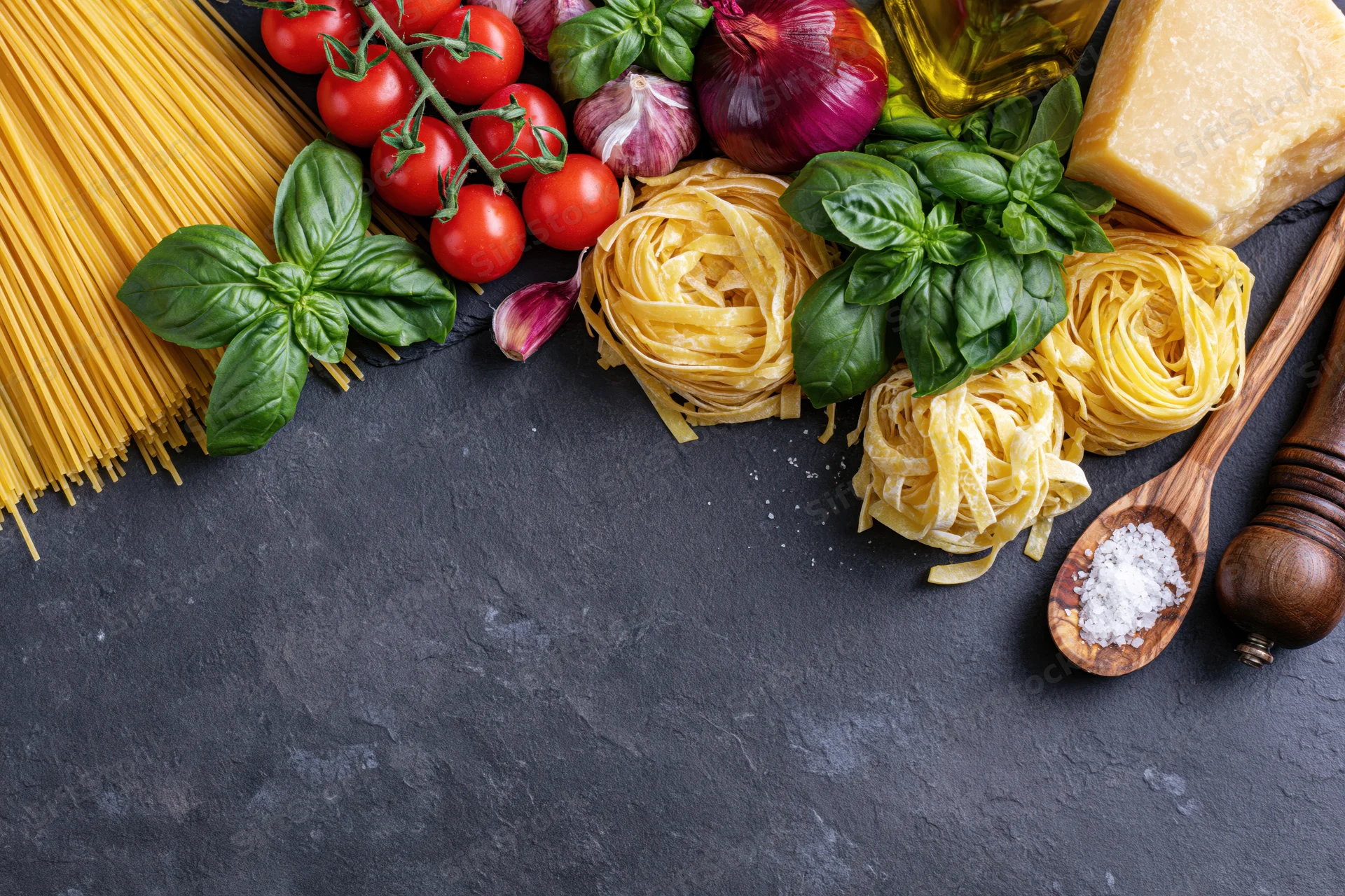 Italian cooking ingredients arranged along the bottom edge of a dark slate surface with copyspace above