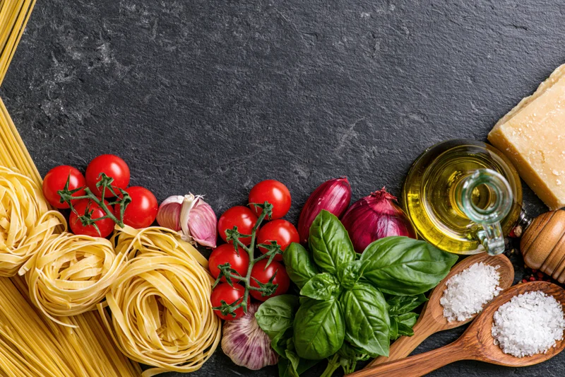 Italian cooking ingredients arranged along the bottom edge of a dark slate surface with copyspace above