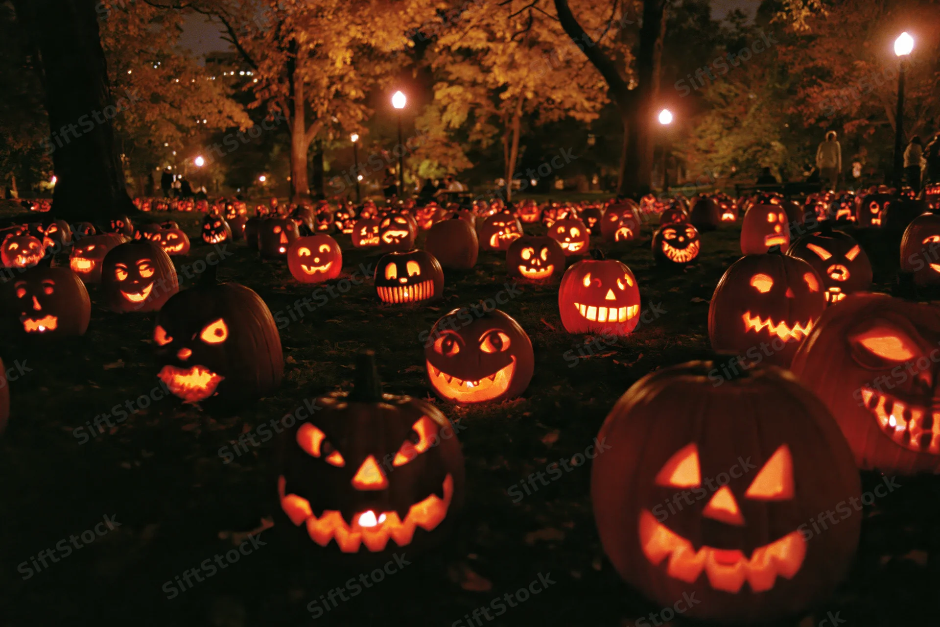Carved jack-o-lanterns glowing in a park at night surrounded by autumn trees