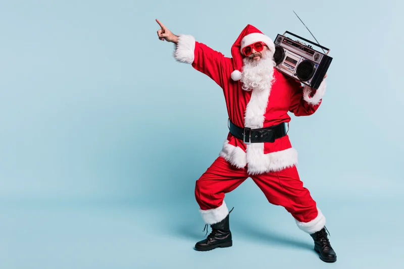 Man dressed as santa claus dancing and pointing while holding a boombox against a light blue background