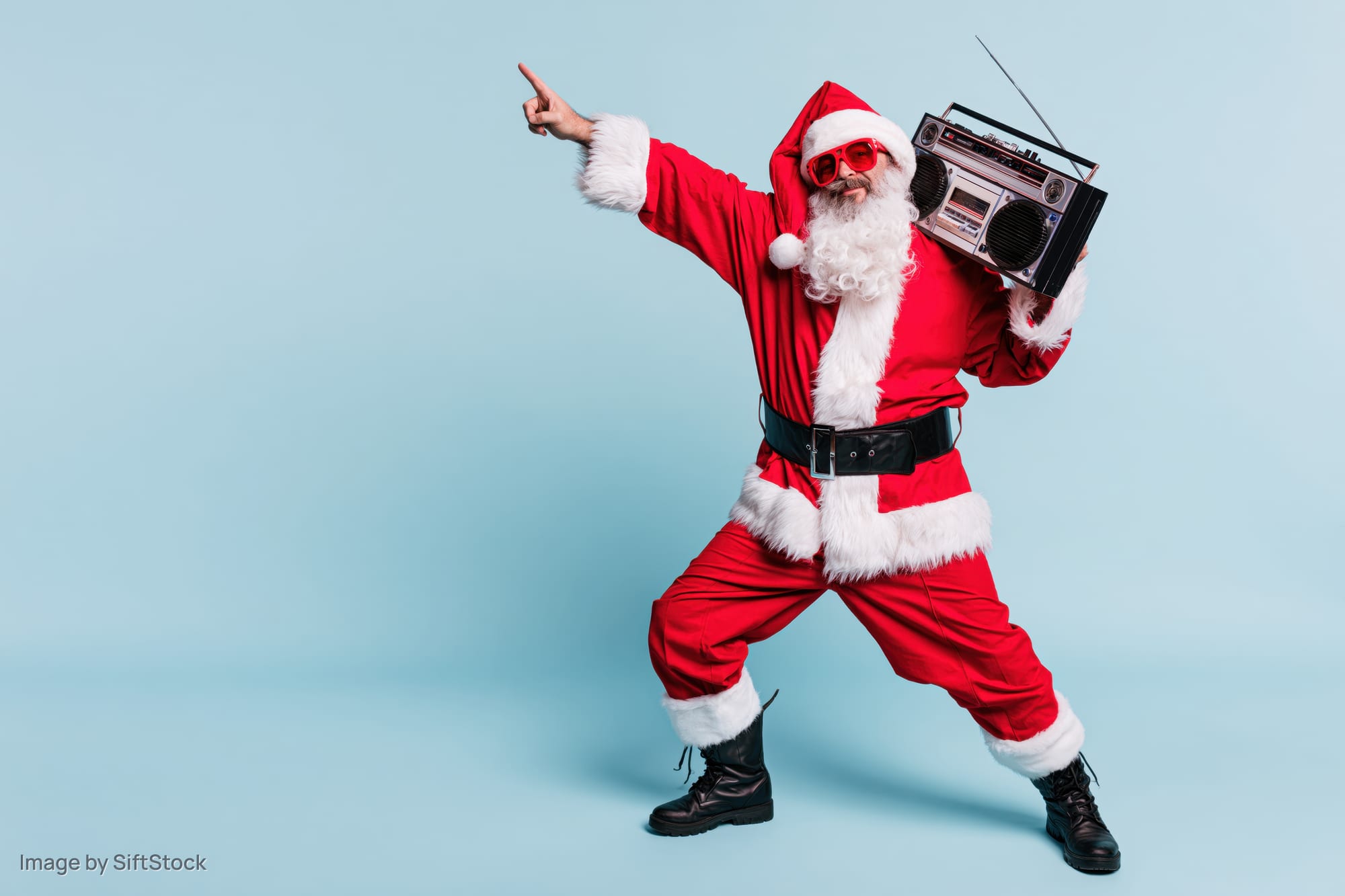 Man dressed as santa claus dancing and pointing while holding a boombox against a light blue background