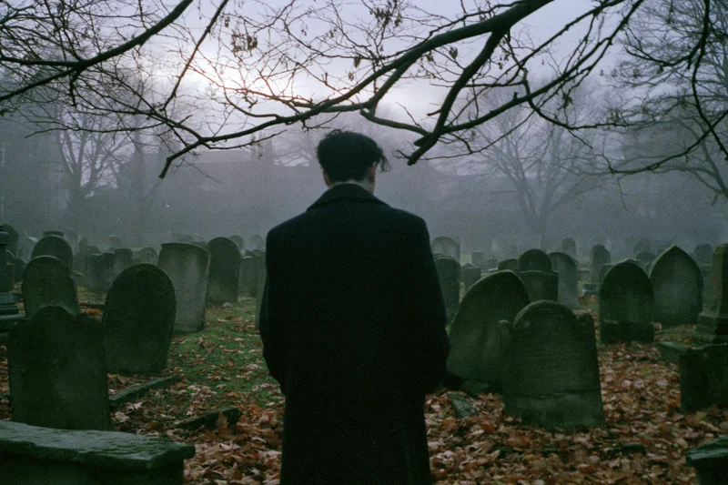 Man in dark overcoat standing with back to camera in foggy cemetery among tombstones and bare trees
